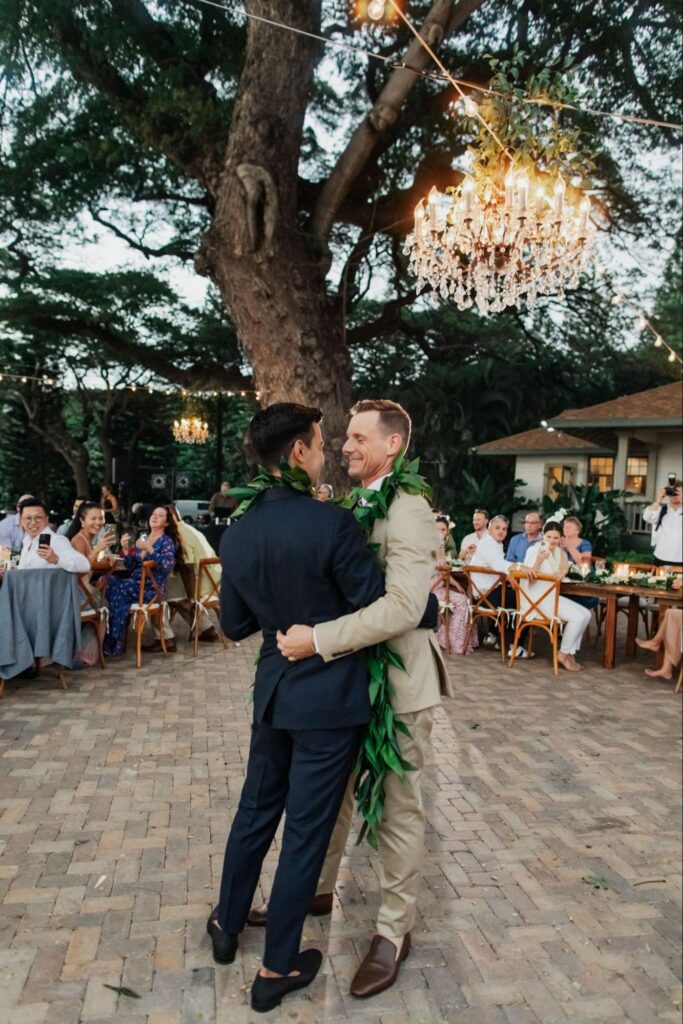 two grooms dancing beneath a chandelier surrounded by friends and family at their Maui wedding