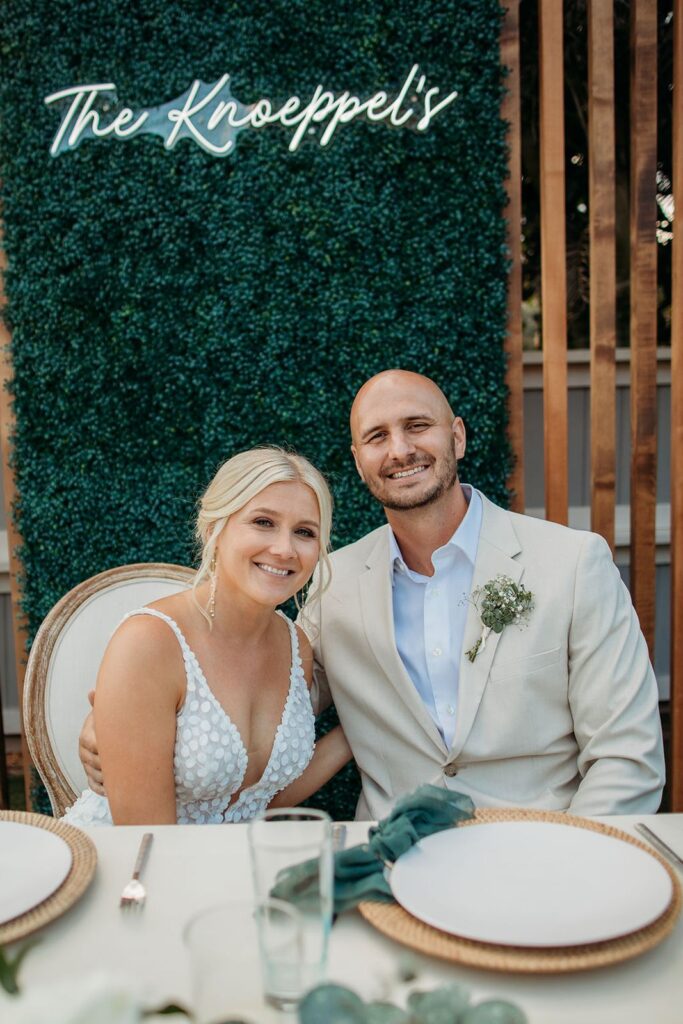 bride and groom seated at sweetheart table with hedge wall and neon sign